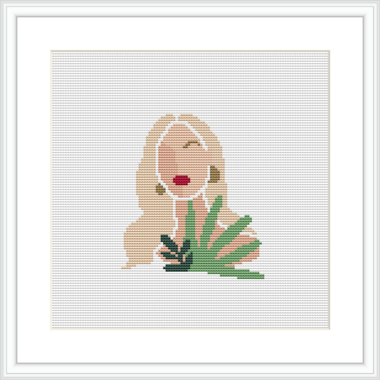 The image depicts a minimalist style cross stitch pattern of a woman with flowing hair and a red lip detail, positioned beside green foliage. The design is displayed within a white frame, placed against a white background.
