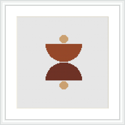 The image shows a framed cross stitch pattern with a central abstract geometric design consisting of a larger, inverted trapezoid stacked upon a smaller one, flanked by two smaller circles, all in various shades of brown on a white background.