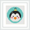 The image shows a cross stitch pattern framed in white. The pattern depicts a roly-poly penguin with a black head, white face, orange beak, and wearing a green circular 'body' suggesting a Christmas bauble. The ornament is topped with a gold cap.