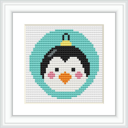 The image shows a cross stitch pattern framed in white. The pattern depicts a roly-poly penguin with a black head, white face, orange beak, and wearing a green circular 'body' suggesting a Christmas bauble. The ornament is topped with a gold cap.