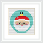 A framed cross stitch pattern of Santa Claus's face depicted within a Christmas ornament. The background is segmented by color, with a dominant green color encircling Santa's face, followed by a white border and a red top suggestive of an ornament cap.