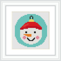 The image depicts a framed cross stitch pattern of a snowman's face with a red and green hat, centered within a circular border. The snowman is smiling and appears on a light grey aida cloth. The frame is white with a rectangular shape and a textured matting around the design.
