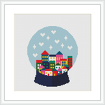 The image depicts a cross stitch pattern of a snow globe containing a colorful village with houses, trees, and snowflakes, all framed within a white border.