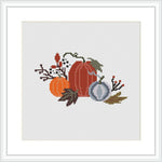 The image shows a cross stitch pattern featuring two pumpkins in shades of orange and gray, surrounded by autumn leaves, berries, and twigs on a white canvas framed in a simple square black frame.