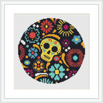 The image showcases a cross stitch pattern with a central skeleton motif surrounded by colorful flowers and geometric patterns, all within an oval-shaped frame on a black background.