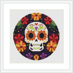 The image depicts a circular cross stitch pattern of a stylized skull surrounded by a border of colorful flowers. It's meant to represent a Mexican-themed Day of the Dead motif.