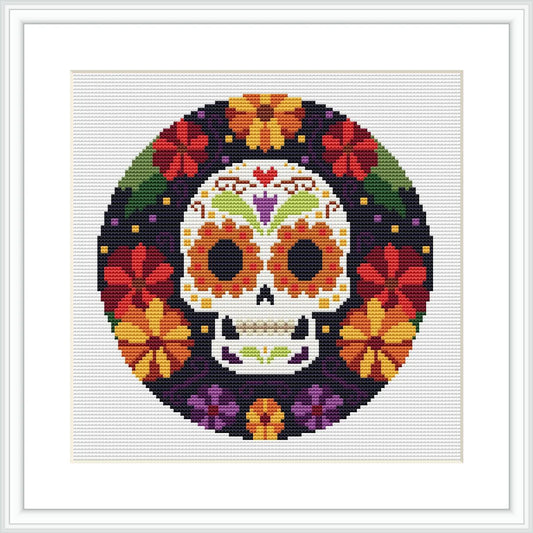 The image depicts a circular cross stitch pattern of a stylized skull surrounded by a border of colorful flowers. It's meant to represent a Mexican-themed Day of the Dead motif.