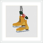 The image depicts a framed cross stitch pattern featuring a pair of hanging ice skates in caramel brown, adorned with green holly leaves and red berries, stitched on a white canvas.