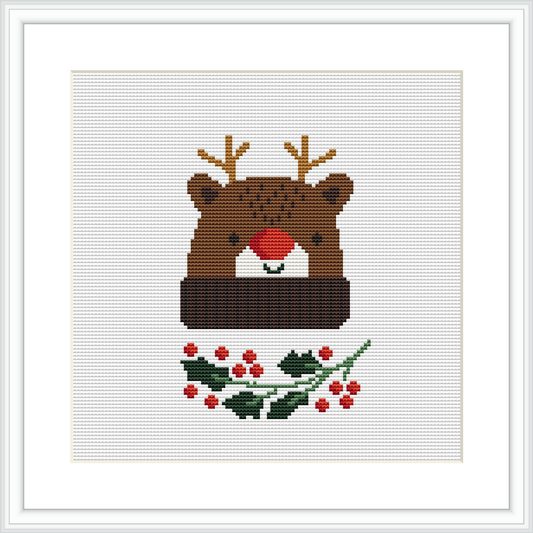 The image features a framed cross stitch pattern of a bear wearing a Christmas hat. The bear is centered on the canvas, surrounded by holly leaves and berries at the bottom. The pattern uses a variety of colors mainly focusing on browns, greens, and reds.