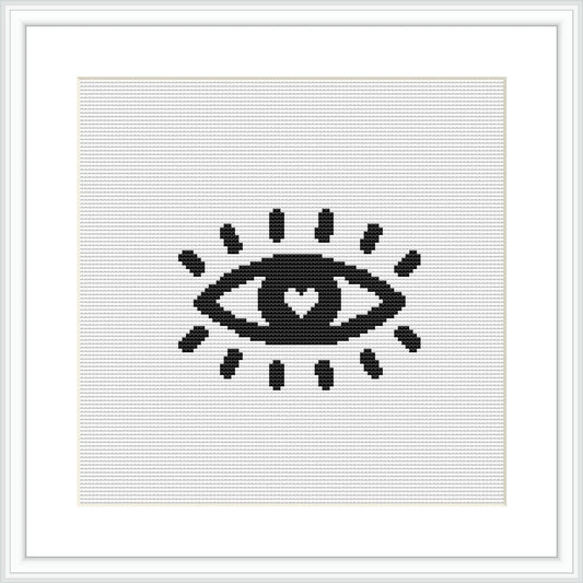 The image shows a monochrome cross-stitch pattern of an eye with a heart in the center, indicating a simple yet profound design, framed white, on a grey background.