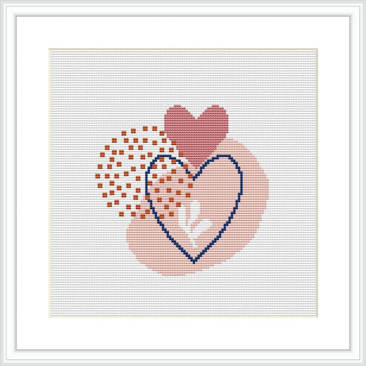 The image shows a framed cross stitch pattern with two overlapping hearts, one filled with a gradient of pink to burgundy, and the other outlined in blue with a peach-toned checkerboard pattern overlapping.