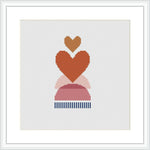 The image features a cross stitch pattern framed in white, depicting stacked abstract hearts in shades of pink, red, and a base with blue stripes, set against a plain white background.