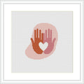 The image displays a framed cross stitch pattern depicting a silhouette of two hands forming a heart shape. The stitches create a gradient effect from reds at the bottom to lighter shades at the top, set against a white background.
