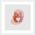 The image displays a framed cross stitch pattern depicting a silhouette of two hands forming a heart shape. The stitches create a gradient effect from reds at the bottom to lighter shades at the top, set against a white background.