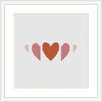 The image shows a cross stitch pattern with three heart shapes in a row, each decreasing in size from left to right. The hearts are filled with different shades of terra cotta and pink, creating a gradient effect. The pattern is displayed within a simulated white frame against a white background.