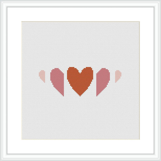 The image shows a cross stitch pattern with three heart shapes in a row, each decreasing in size from left to right. The hearts are filled with different shades of terra cotta and pink, creating a gradient effect. The pattern is displayed within a simulated white frame against a white background.