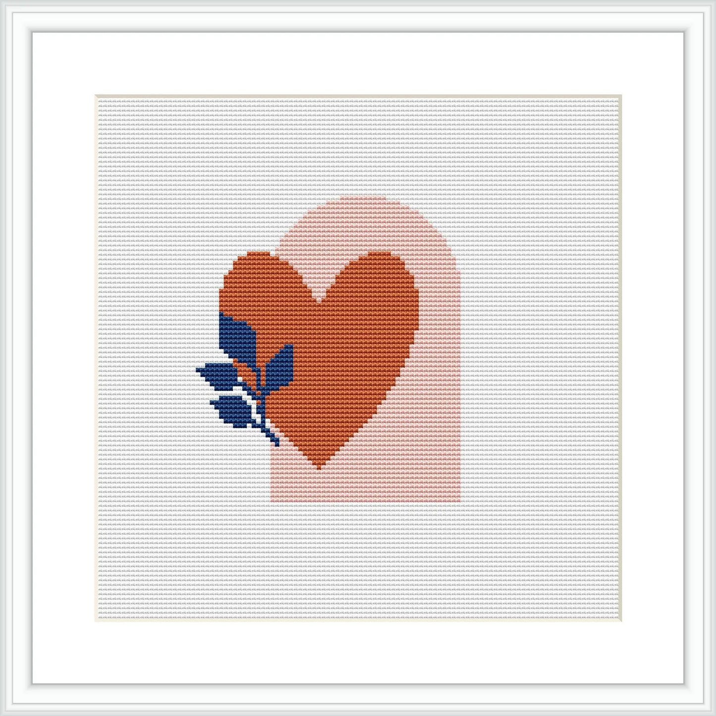 The image shows a framed cross stitch pattern of a terracotta heart with a small blue and green botanical element on the right side, on a pastel pink background bordered by white.