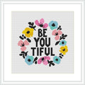 The image shows a cross stitch pattern featuring the phrase 'BE YOU TIFUL' in block letters, encircled by a colorful floral wreath. The flowers are pink, blue, and yellow, and the text is in black against a white background. The design is framed within a simple square frame.