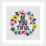 The image shows a cross stitch pattern featuring the phrase 'BE YOU TIFUL' in block letters, encircled by a colorful floral wreath. The flowers are pink, blue, and yellow, and the text is in black against a white background. The design is framed within a simple square frame.