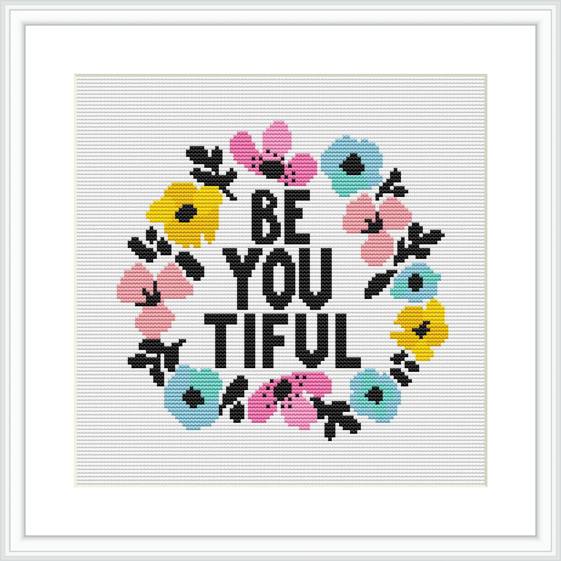 The image shows a cross stitch pattern featuring the phrase 'BE YOU TIFUL' in block letters, encircled by a colorful floral wreath. The flowers are pink, blue, and yellow, and the text is in black against a white background. The design is framed within a simple square frame.