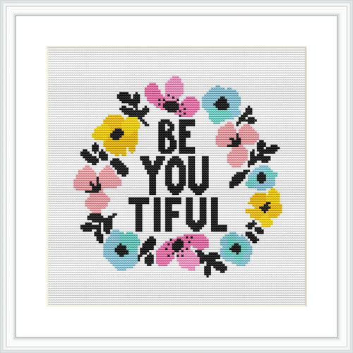 The image shows a cross stitch pattern featuring the phrase 'BE YOU TIFUL' in block letters, encircled by a colorful floral wreath. The flowers are pink, blue, and yellow, and the text is in black against a white background. The design is framed within a simple square frame.