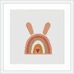 The image shows a framed cross stitch embroidery of a stylized bunny head silhouette with its ears extending upwards. The silhouette is patterned with rainbow stripes in earthy tones, set against a white background within the frame.