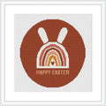 The image displays a framed cross stitch pattern showing a stylized brown bunny above a rainbow arc with the phrase 'HAPPY EASTER' at the bottom, all contained within a circular border.