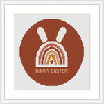 The image displays a framed cross stitch pattern showing a stylized brown bunny above a rainbow arc with the phrase 'HAPPY EASTER' at the bottom, all contained within a circular border.