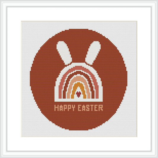 The image displays a framed cross stitch pattern showing a stylized brown bunny above a rainbow arc with the phrase 'HAPPY EASTER' at the bottom, all contained within a circular border.