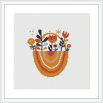 The image showcases a framed cross stitch pattern featuring a stylized bunny in the center of an arching rainbow, with assorted flowers and plants adorning the rainbow's curves, all stitched on a white fabric.