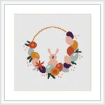 The cross stitch design features a central pink bunny flanked by an arc composed of Easter eggs and flowers. The eggs and flowers are rendered in shades of purple, orange, and green, with scattered pink blossoms creating a festive wreath-like appearance against a white background.