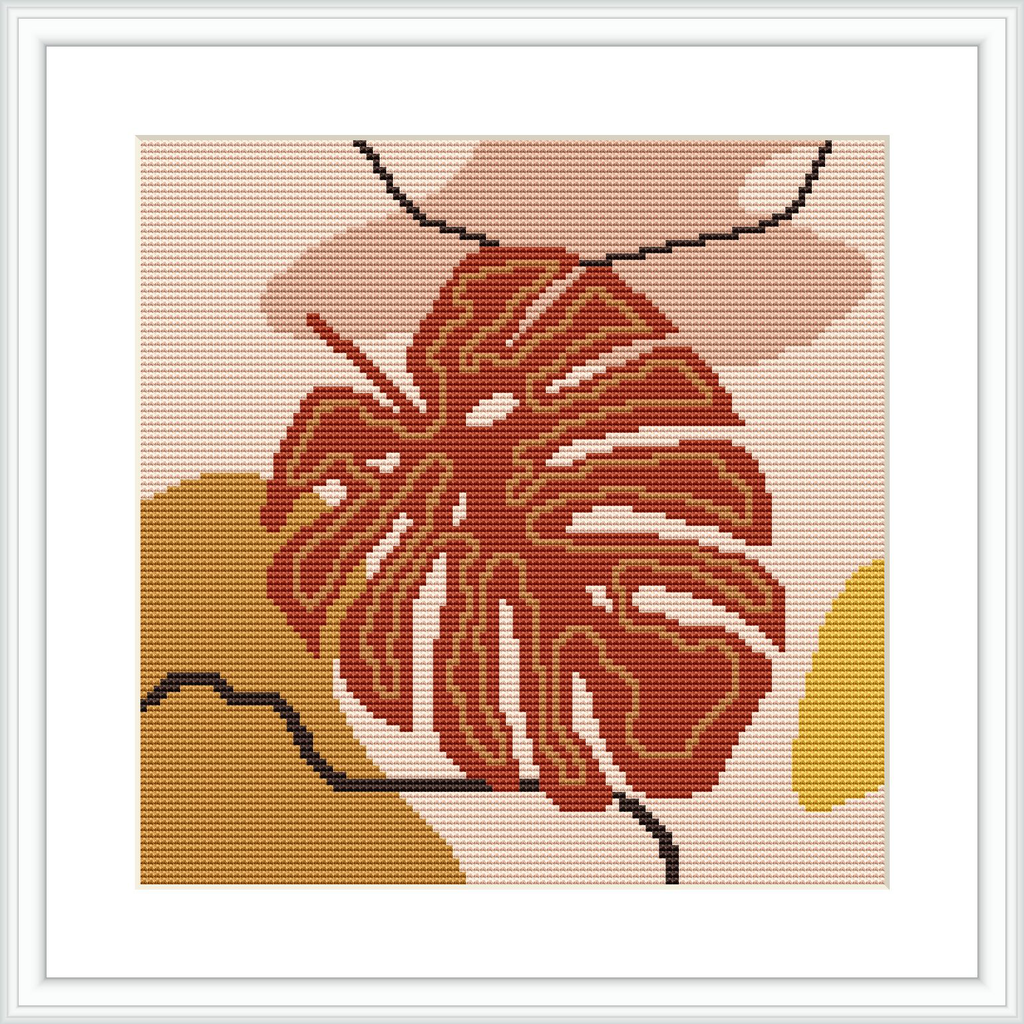 A framed cross stitch pattern of a monstera leaf in earthy tones, with terracotta, beige, and cream colors. The pattern appears to be set against a light-colored background and framed in a simple white frame.