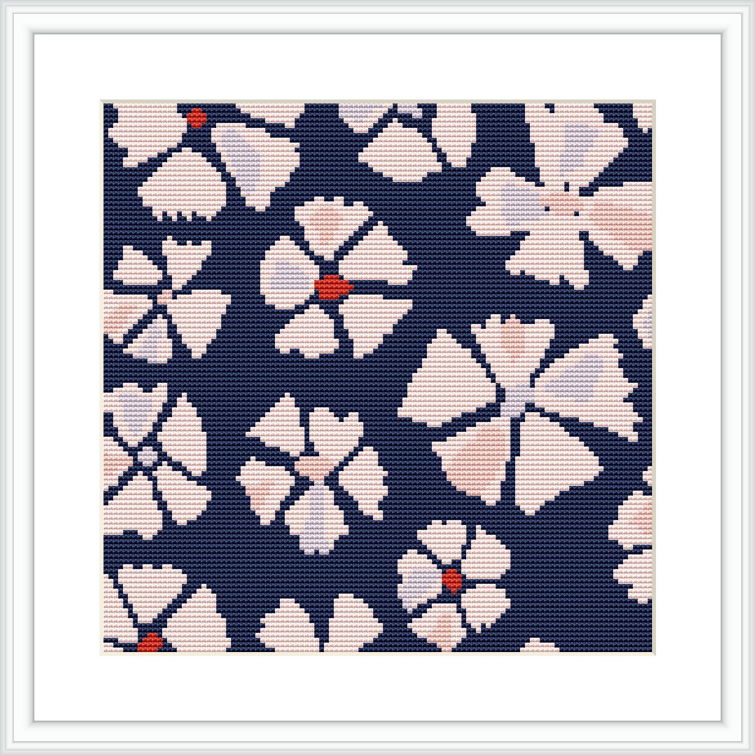 The image displays a framed cross stitch pattern with a design of pink flowers on a dark blue background. Each flower features a central orange accent and is surrounded by smaller white and cream details.