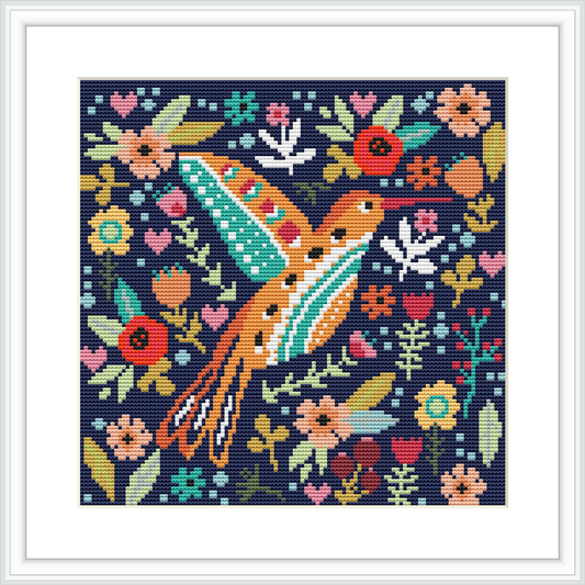 The image shows a framed square cross stitch pattern. In the center, a blue hummingbird hovers, surrounded by a variety of colorful flowers and leaves against a dark background.