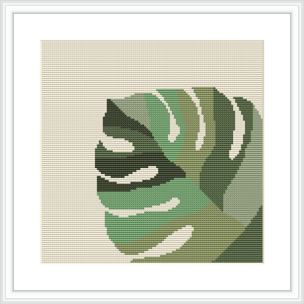 The image displays a framed cross stitch pattern of a monstera leaf with various shades of green and some white highlights on a beige background, within a light-coloured square frame on a wall.
