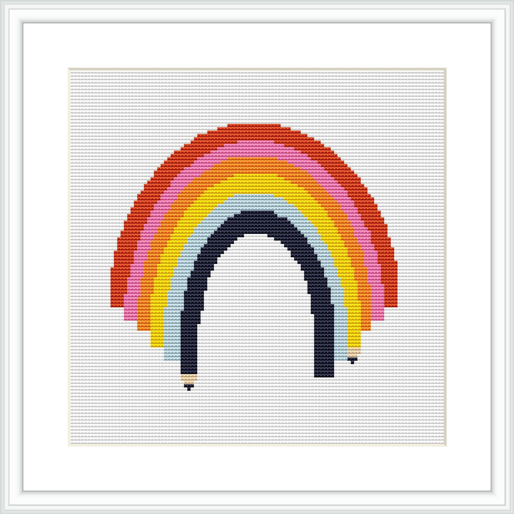 The image showcases a framed cross stitch pattern of a rainbow with seven arching bands of color on a white background.