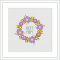 The image depicts a cross-stitch pattern with a circular floral wreath design. In the center, the words 'BEST MOM' are written in capital letters. The flowers are primarily pink and purple with green leaves and yellow accents, arranged symmetrically on a plain white background.