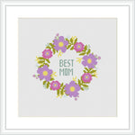 The image depicts a cross-stitch pattern with a circular floral wreath design. In the center, the words 'BEST MOM' are written in capital letters. The flowers are primarily pink and purple with green leaves and yellow accents, arranged symmetrically on a plain white background.