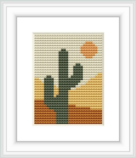 The image showcases a cross stitch pattern framed in white, portraying a stylized desert scene with a prominent cactus in the foreground against a backdrop of rolling dunes and a round sun hanging in the sky.