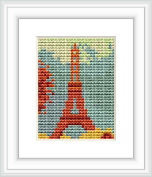 The cross stitch depicts the Eiffel Tower centered on the canvas, with a splash of abstract floral element to the left, set against a backdrop of sky blue, all framed with a white border.