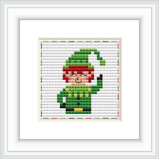 The image showcases a framed cross-stitch pattern of a cartoonish elf. The elf is primarily green with red and white accents, set against a patterned background.