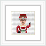 The image depicts a framed cross-stitch pattern of Mrs. Claus with glasses, wearing a red outfit with a green background on a wall.