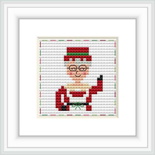 The image depicts a framed cross-stitch pattern of Mrs. Claus with glasses, wearing a red outfit with a green background on a wall.