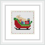 The image shows a framed cross stitch pattern of Santa's sleigh filled with colorful gift boxes. The sleigh is predominantly red and green, set against a snowy background.