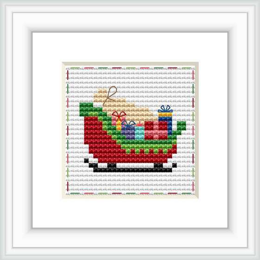 The image shows a framed cross stitch pattern of Santa's sleigh filled with colorful gift boxes. The sleigh is predominantly red and green, set against a snowy background.
