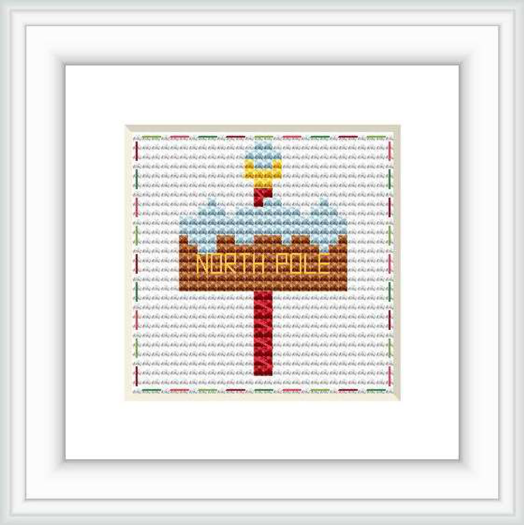 The image displays a cross stitch pattern with a festive North Pole signpost. The sign is centered on a white background with 'NORTH POLE' in golden letters. It is adorned with a red arrow and small Christmas decorations.