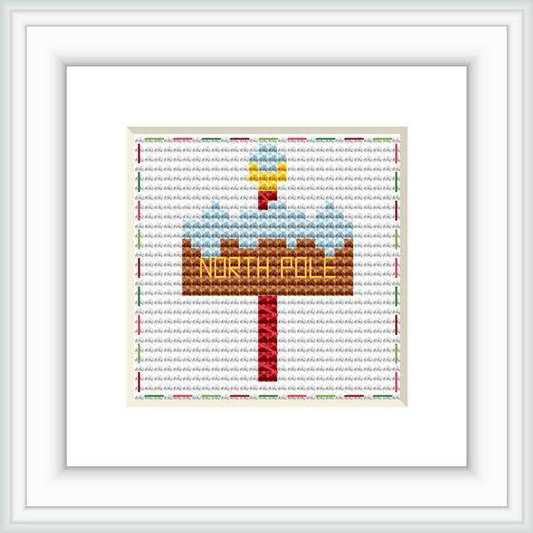 The image displays a cross stitch pattern with a festive North Pole signpost. The sign is centered on a white background with 'NORTH POLE' in golden letters. It is adorned with a red arrow and small Christmas decorations.