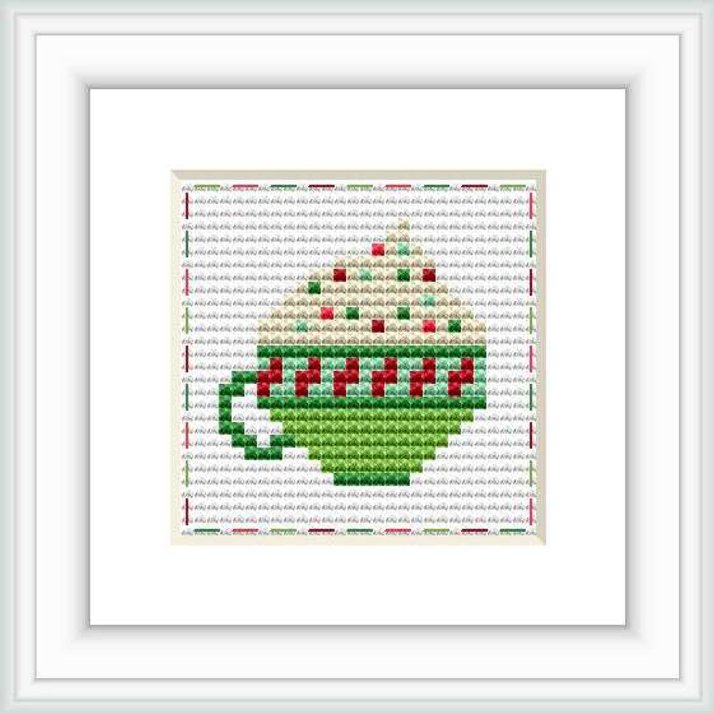 The image displays a square cross stitch pattern with a green and red mug depicted in the center. The mug has a heart design and is surrounded by a border of cross-stitched hearts on a white background. It is matted in a frame for presentation.