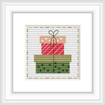 The image shows a cross stitch pattern framed in white, depicting a stylized gift box with a bow on top. The pattern is set against a patterned background with faint script text. The gift box has diagonal red and white stripes, and the bow appears to be a darker shade of green.