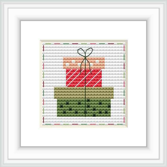 The image shows a cross stitch pattern framed in white, depicting a stylized gift box with a bow on top. The pattern is set against a patterned background with faint script text. The gift box has diagonal red and white stripes, and the bow appears to be a darker shade of green.